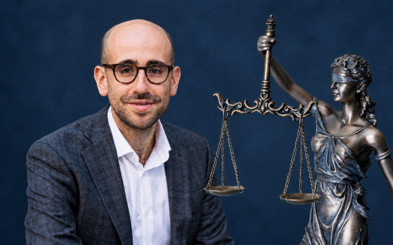 Jacob Abolafia in glasses and a blazer beside a bronze statue of blindfolded Lady Justice holding scales, against a dark blue background